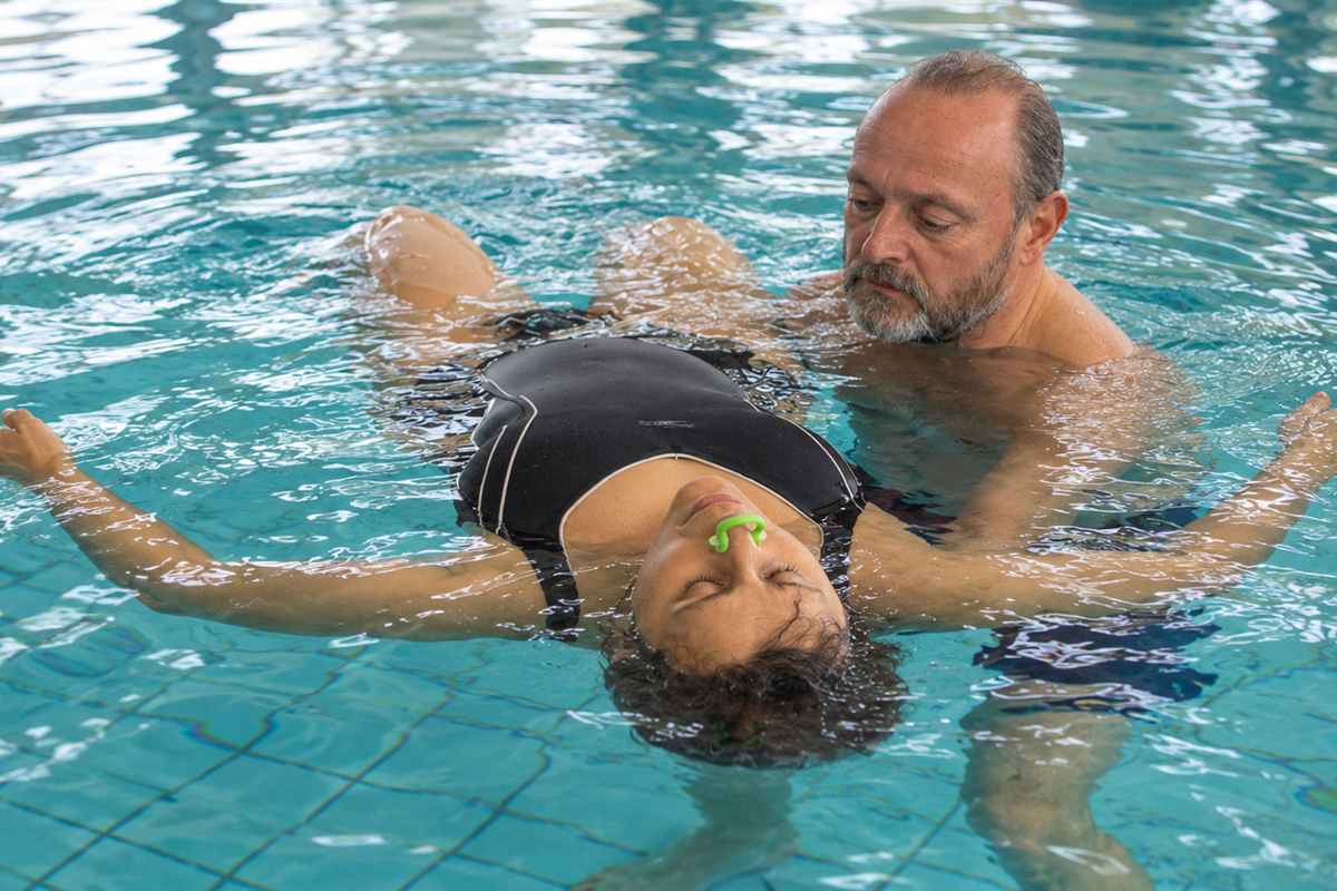 Coccoloterapia® — Paolo Cericola in acqua Paolo Cericola sostiene una donna che galleggia supina, con le braccia aperte e uno stringinaso verde.