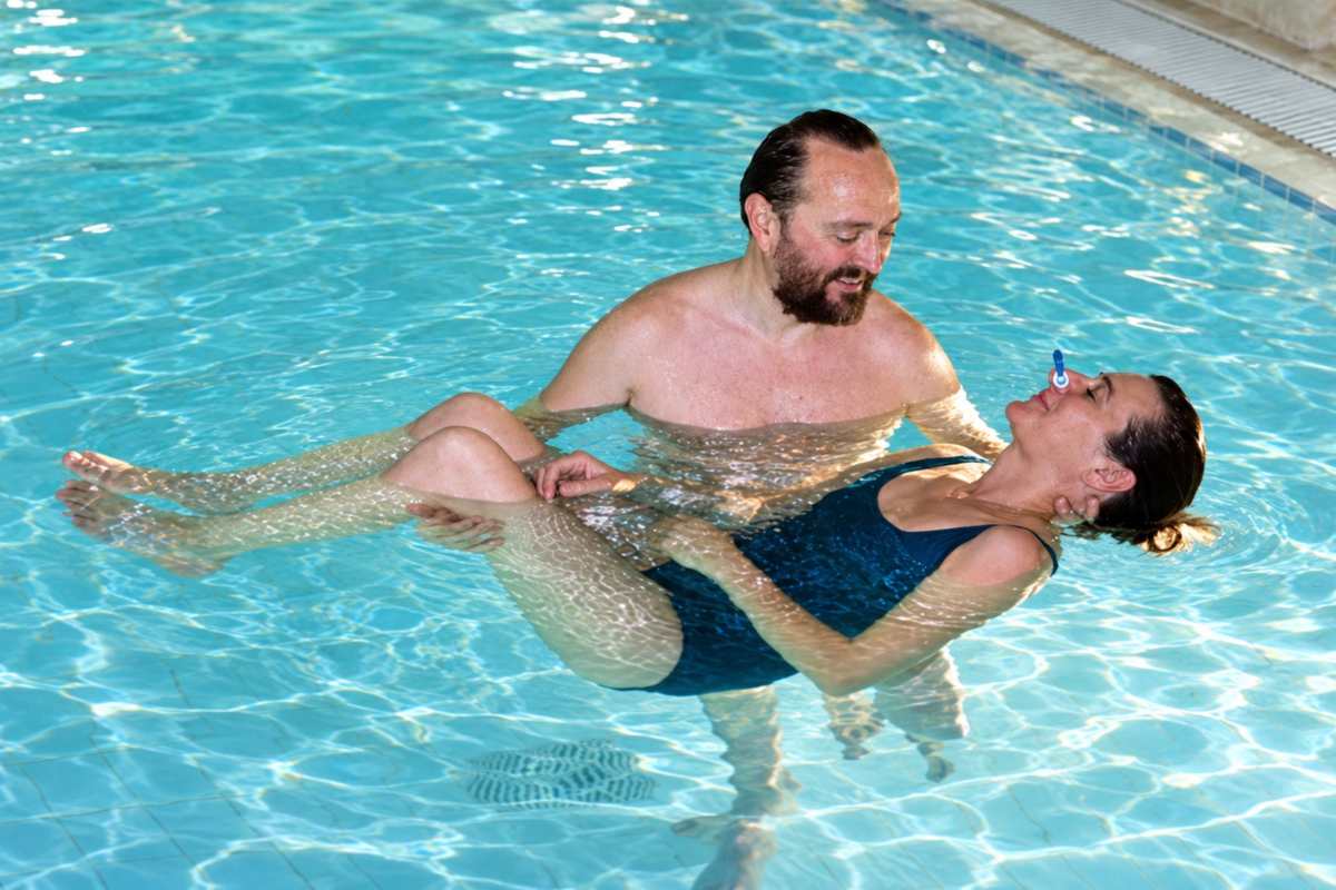 Paolo Cericola – Coccoloterapia® in acqua calda Paolo Cericola sostiene una donna che galleggia sulla schiena con le ginocchia piegate e gli occhi chiusi in piscina
